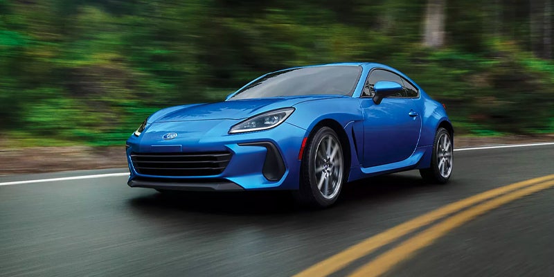 A blue Subaru BRZ sports car driving on a winding road with trees in the background.
