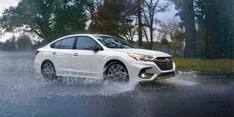White Subaru Legacy Sedan drives through a large puddle, splashing water onto the road.