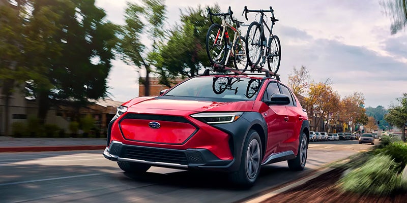 Red Subaru Solterra SUV with two bicycles mounted on its roof rack, driving down a tree-lined street.